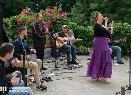 fotografia. dni wyszehradzkie w willi decjusza. koncert zespołu balkan artz. kobieta w długiej fioletowej spódnicy śpiewa. za nią na kamiennym tarasie willi w półkolu siedzą pozostali członkowie zespołu z instrumentami.